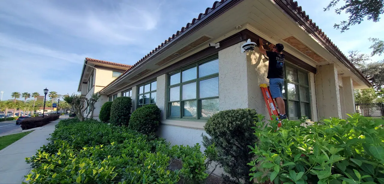 Technician on a ladder mounting a camera under a tile-roof eave